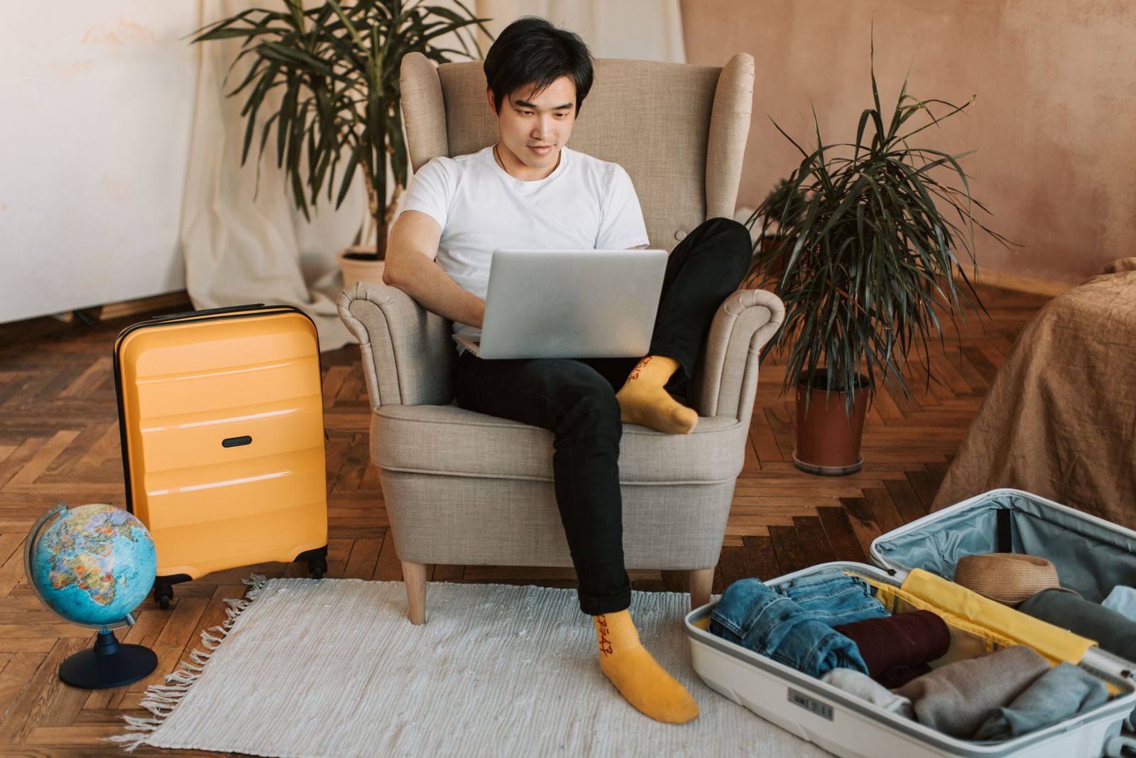 Man in an armchair checking a Carnival cruise price guarantee on a laptop beside an open suitcase and globe
