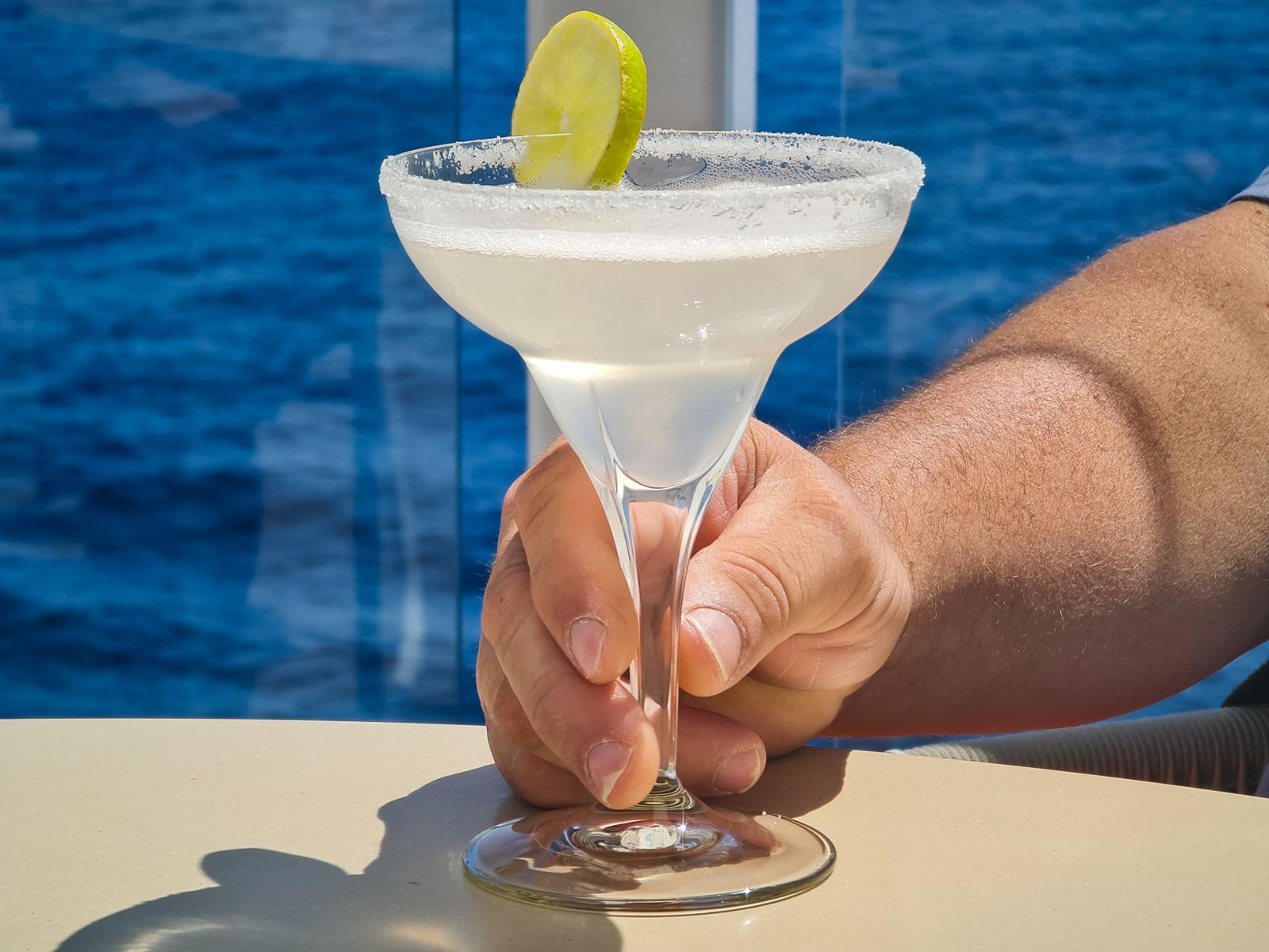 Hand holding a margarita with a lime slice on a ship deck above the blue sea, the kind of extra a Princess cruise price guarantee credit pays for
